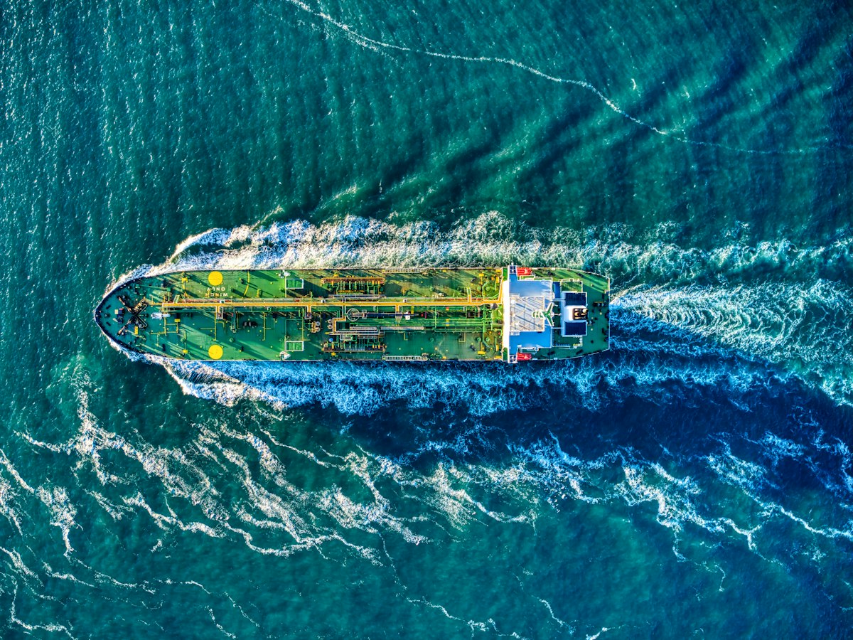 A green tanker seen from above, cutting a white wake through teal water